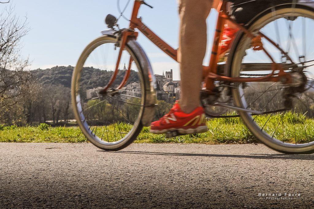 Une personne roulant sur un vélo orange sur un chemin ensoleillé, portant des chaussures rouge vif, près du gîte du Mas Saint Antoine en Ardèche, avec un village et une église en arrière-plan flou. L’herbe verte et les arbres bordent cette scène paisible. Une personne roulant sur un vélo orange sur un chemin ensoleillé, portant des chaussures rouge vif, près du gîte du Mas Saint Antoine en Ardèche, avec un village et une église en arrière-plan flou. L'herbe verte et les arbres bordent cette scène paisible.