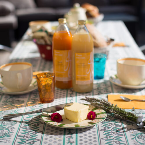 Une table extérieure ensoleillée pour le petit-déjeuner au Mas Saint Antoine, avec deux bouteilles de jus, des mugs, des verres colorés, des assiettes avec du beurre et des garnitures, et un couteau. La nappe à feuilles ajoute une touche douillette à cet hébergement Ardèche méridionale pour les séjours en groupe.