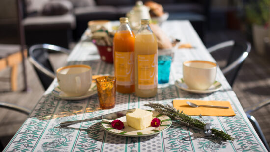 Une table extérieure ensoleillée pour le petit-déjeuner au Mas Saint Antoine, avec deux bouteilles de jus, des mugs, des verres colorés, des assiettes avec du beurre et des garnitures, et un couteau. La nappe à feuilles ajoute une touche douillette à cet hébergement Ardèche méridionale pour les séjours en groupe.