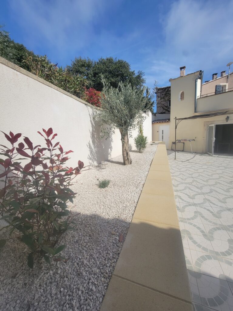 Cour méditerranéenne ensoleillée au Mas Saint Antoine avec jardin de gravier blanc, olivier, arbuste à feuilles rouges, terrasse carrelée à motifs, et maison de gîte ardéchoise de couleur crème sous un ciel bleu.