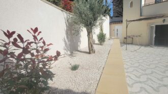 Cour méditerranéenne ensoleillée au Mas Saint Antoine avec jardin de gravier blanc, olivier, arbuste à feuilles rouges, terrasse carrelée à motifs, et maison de gîte ardéchoise de couleur crème sous un ciel bleu.