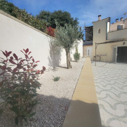Cour méditerranéenne ensoleillée au Mas Saint Antoine avec jardin de gravier blanc, olivier, arbuste à feuilles rouges, terrasse carrelée à motifs, et maison de gîte ardéchoise de couleur crème sous un ciel bleu.