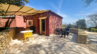 Une petite maison rouge au Mas Saint Antoine avec un patio ombragé comprenant une chaise jaune, une table blanche, plusieurs chaises noires et un accès à une piscine, le tout entouré de murs en pierre et d'arbres sous le ciel bleu clair de l'hébergement Ardèche méridionale.