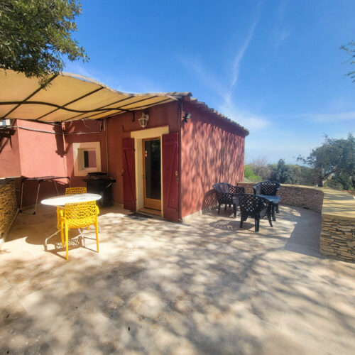 Une petite maison rouge au Mas Saint Antoine avec un patio ombragé comprenant une chaise jaune, une table blanche, plusieurs chaises noires et un accès à une piscine, le tout entouré de murs en pierre et d'arbres sous le ciel bleu clair de l'hébergement Ardèche méridionale.