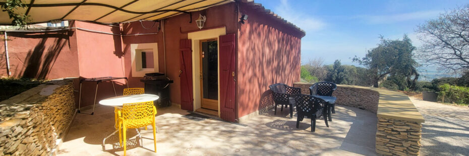 Une petite maison rouge au Mas Saint Antoine avec un patio ombragé comprenant une chaise jaune, une table blanche, plusieurs chaises noires et un accès à une piscine, le tout entouré de murs en pierre et d'arbres sous le ciel bleu clair de l'hébergement Ardèche méridionale.