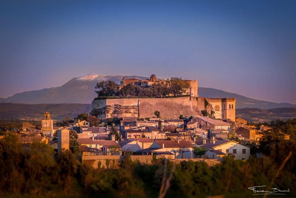 Un château perché aux murs de pierre surplombe une ville aux bâtiments blancs au coucher du soleil dans l’Ardèche méridionale, avec des montagnes lointaines et une lumière dorée illuminant un gîte tranquille de l’Ardèche et une piscine étincelante en contrebas. Vue du château de Grignan dominant le village médiéval, Drôme provençale, à 30 minutes du Mas Saint Antoine
