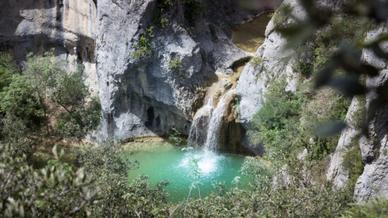 Une petite cascade se jette dans un bassin turquoise entouré de falaises rocheuses et d'une végétation luxuriante, vue d'en haut à travers des branches en surplomb près du Mas Saint Antoine, un gîte accueillant en Ardèche parfait pour votre escapade en groupe.