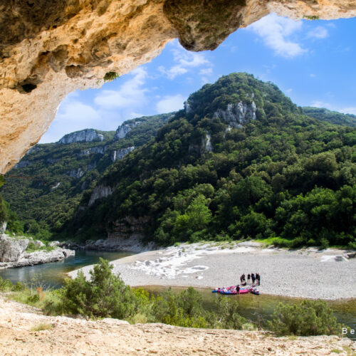 Depuis l'intérieur d'une grotte rocheuse, on aperçoit une rivière qui serpente à travers des collines verdoyantes et boisées près du gîte Ardèche Mas Saint Antoine. Sur une rive sablonneuse en contrebas, des gens se tiennent près de kayaks aux couleurs vives sous un ciel partiellement nuageux.