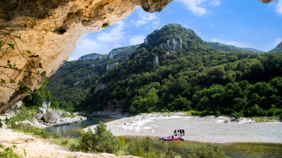 Depuis l'intérieur d'une grotte rocheuse, on aperçoit une rivière qui serpente à travers des collines verdoyantes et boisées près du gîte Ardèche Mas Saint Antoine. Sur une rive sablonneuse en contrebas, des gens se tiennent près de kayaks aux couleurs vives sous un ciel partiellement nuageux.