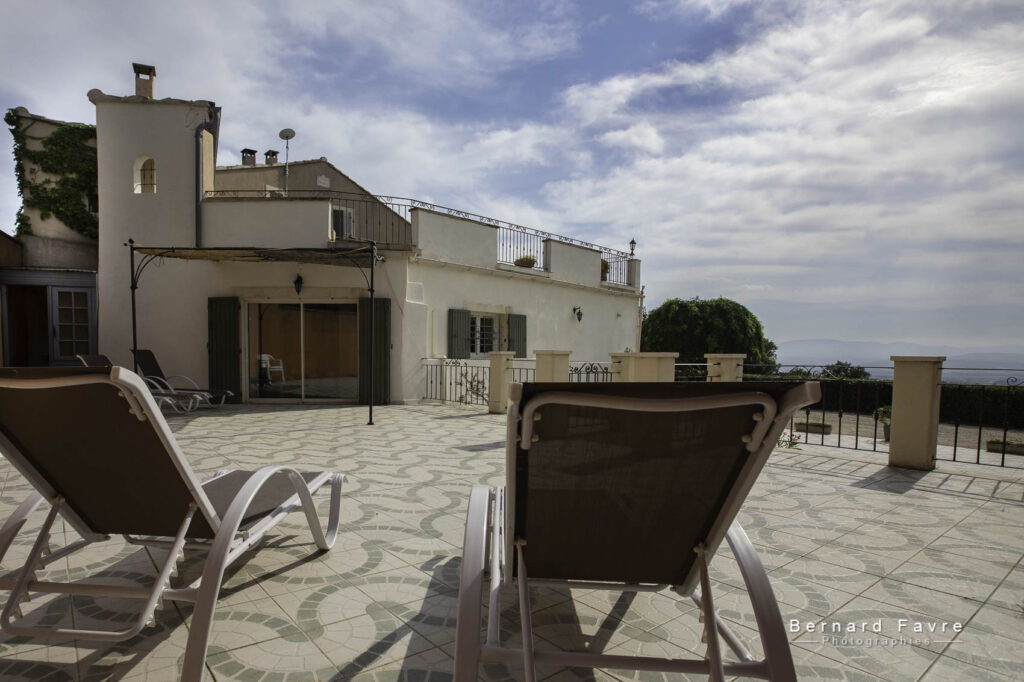 Deux chaises longues font face à une terrasse carrelée spacieuse, avec un gîte blanc de style méditerranéen en arrière-plan, surplombant un paysage pittoresque, une piscine et un ciel bleu partiellement nuageux.