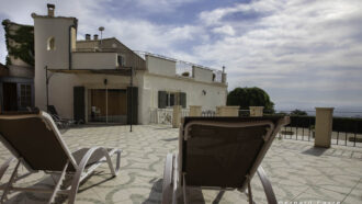 Deux chaises longues font face à une terrasse carrelée spacieuse, avec un gîte blanc de style méditerranéen en arrière-plan, surplombant un paysage pittoresque, une piscine et un ciel bleu partiellement nuageux.