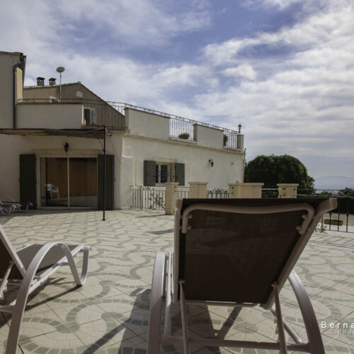 Deux chaises longues font face à une terrasse carrelée spacieuse, avec un gîte blanc de style méditerranéen en arrière-plan, surplombant un paysage pittoresque, une piscine et un ciel bleu partiellement nuageux.