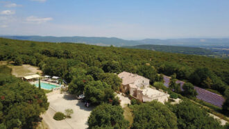 Vue aérienne du Mas Saint Antoine, gîte de charme ardéchois pour les séjours en groupe, entouré d'arbres, d'une piscine à gauche, de champs de lavande à droite, et de collines ondulantes en arrière-plan sous un ciel bleu clair.