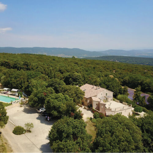 Vue aérienne du Mas Saint Antoine, gîte de charme ardéchois pour les séjours en groupe, entouré d'arbres, d'une piscine à gauche, de champs de lavande à droite, et de collines ondulantes en arrière-plan sous un ciel bleu clair.