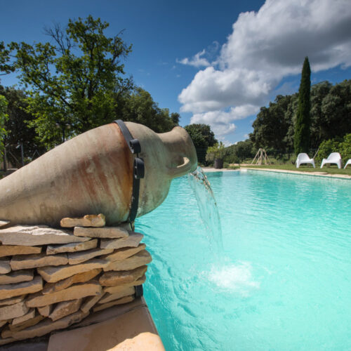 Une grande urne en terre cuite inclinée sur le côté verse de l'eau dans une piscine bleu vif au Mas Saint Antoine, entourée de verdure et de pierres, avec des chaises longues blanches à l'arrière-plan sous un ciel partiellement nuageux.