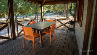 Un porche en bois au Mas Saint Antoine avec une table et quatre chaises orange vif, entouré d'arbres et de soleil. Un petit grill se trouve près de la balustrade, avec un chemin de gravier à proximité - un hébergement Ardèche méridionale parfait pour votre séjour.