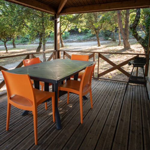 Un porche en bois au Mas Saint Antoine avec une table et quatre chaises orange vif, entouré d'arbres et de soleil. Un petit grill se trouve près de la balustrade, avec un chemin de gravier à proximité - un hébergement Ardèche méridionale parfait pour votre séjour.