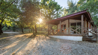 Une petite cabane en bois avec un porche se trouve au milieu des arbres, la lumière du soleil passant à travers les branches, idéale pour les séjours en groupe ou comme gîte en Ardèche, projetant de longues ombres sur une allée en gravier et sur le sol forestier environnant.