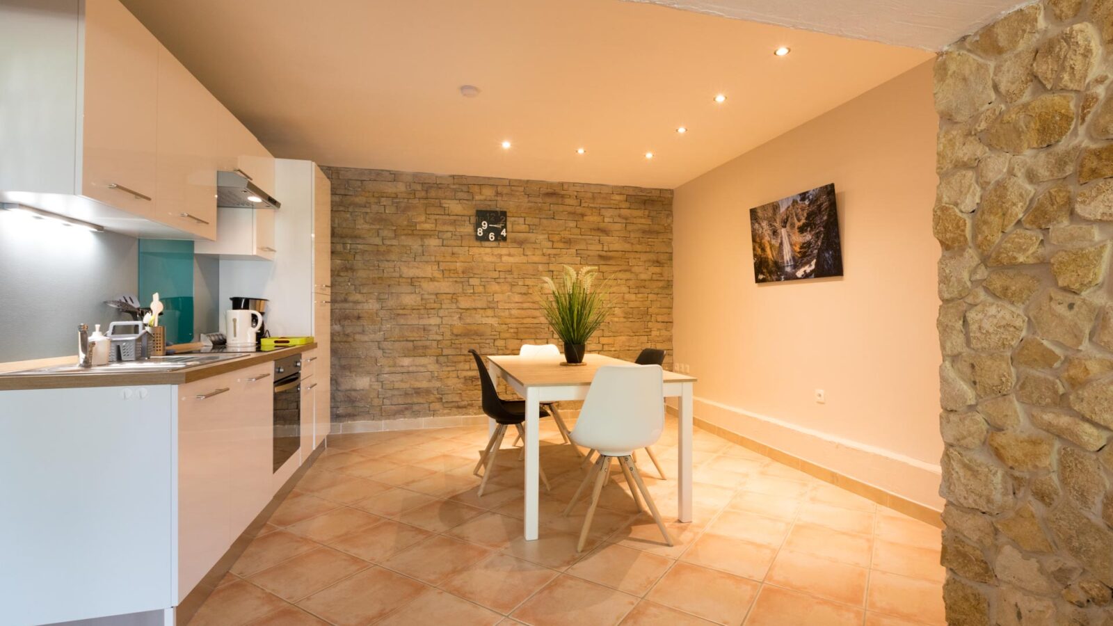 Modern kitchen and dining area with white cabinets, built-in appliances, a wooden table with four chairs, and a potted plant centerpiece evoke the warmth of a charming cottage in Ardèche, France. Stone accent wall and tile flooring add rustic appeal. Modern kitchen and dining area with white cabinets, built-in appliances, a wooden table with four chairs, and a potted plant centerpiece evoke the warmth of a charming cottage in Ardèche, France. Stone accent wall and tile flooring add rustic appeal.