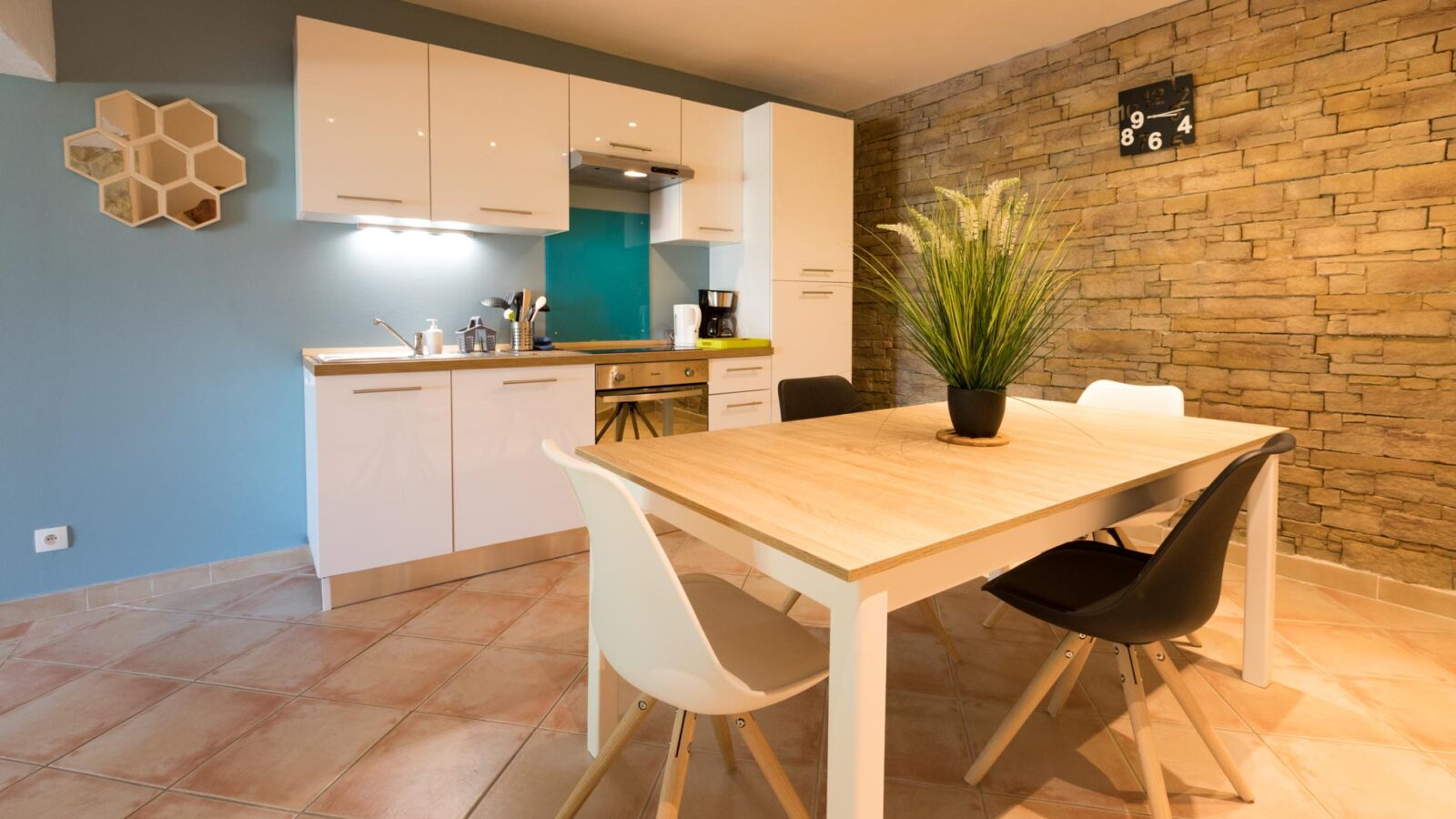 Modern kitchen and dining area with light tiled floors, white cabinets, a blue accent wall, and a stone feature wall. Ideal for a groupe stay at Mas Saint Antoine, the space features a wooden dining table with four chairs and a potted plant. Modern kitchen and dining area with light tiled floors, white cabinets, a blue accent wall, and a stone feature wall. Ideal for a groupe stay at Mas Saint Antoine, the space features a wooden dining table with four chairs and a potted plant.