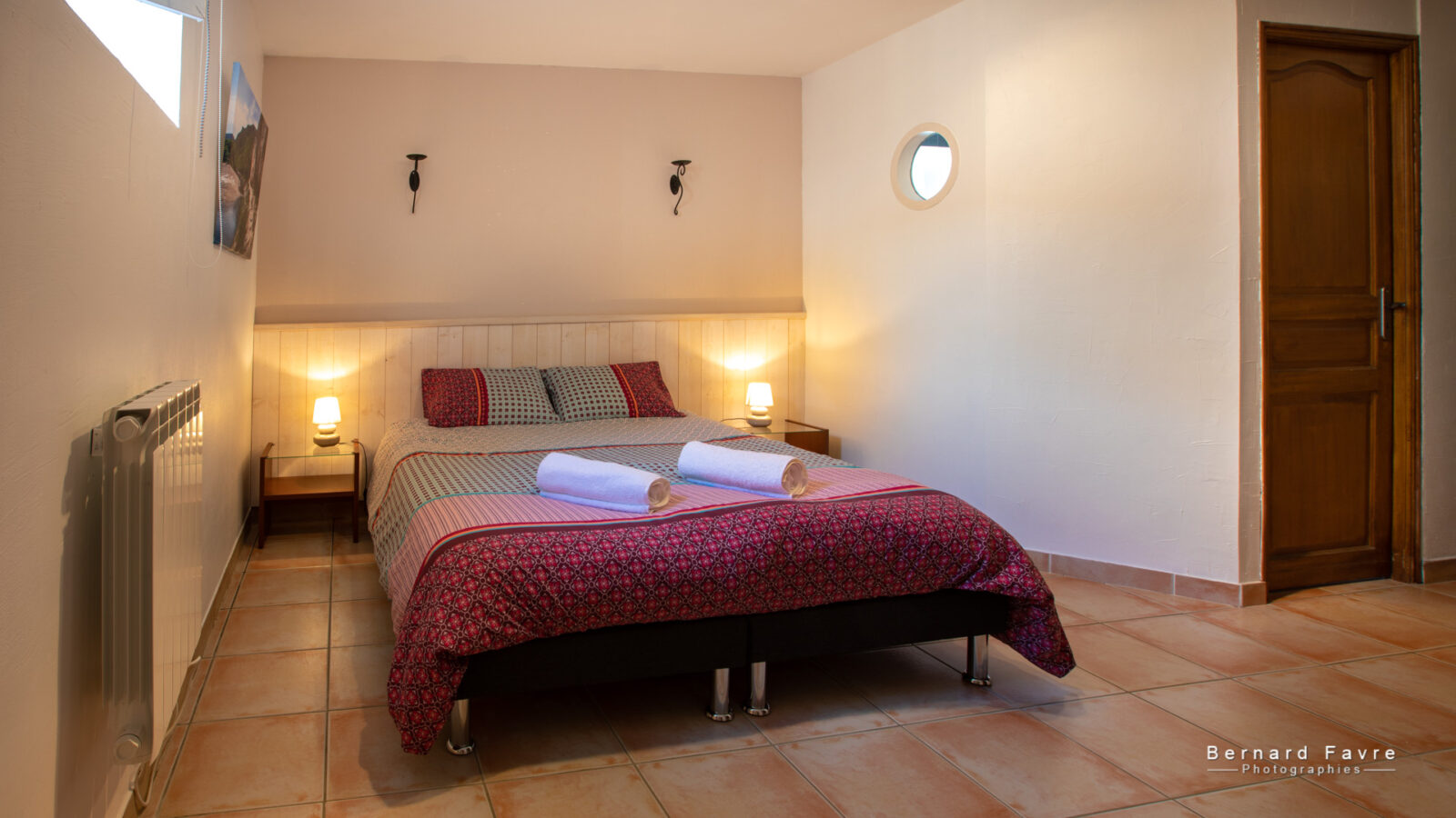 Un gîte confortable 4 personnes Ardèche avec piscine chambre avec un lit double recouvert d'une literie à motifs, deux serviettes roulées sur le dessus, tables de chevet avec lampes, sol carrelé, une porte en bois, et un éclairage doux. Un gîte confortable 4 personnes Ardèche avec piscine chambre avec un lit double recouvert d'une literie à motifs, deux serviettes roulées sur le dessus, tables de chevet avec lampes, sol carrelé, une porte en bois, et un éclairage doux.