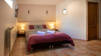 Un gîte confortable 4 personnes Ardèche avec piscine chambre avec un lit double recouvert d'une literie à motifs, deux serviettes roulées sur le dessus, tables de chevet avec lampes, sol carrelé, une porte en bois, et un éclairage doux.