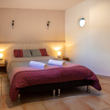Un gîte confortable 4 personnes Ardèche avec piscine chambre avec un lit double recouvert d'une literie à motifs, deux serviettes roulées sur le dessus, tables de chevet avec lampes, sol carrelé, une porte en bois, et un éclairage doux.