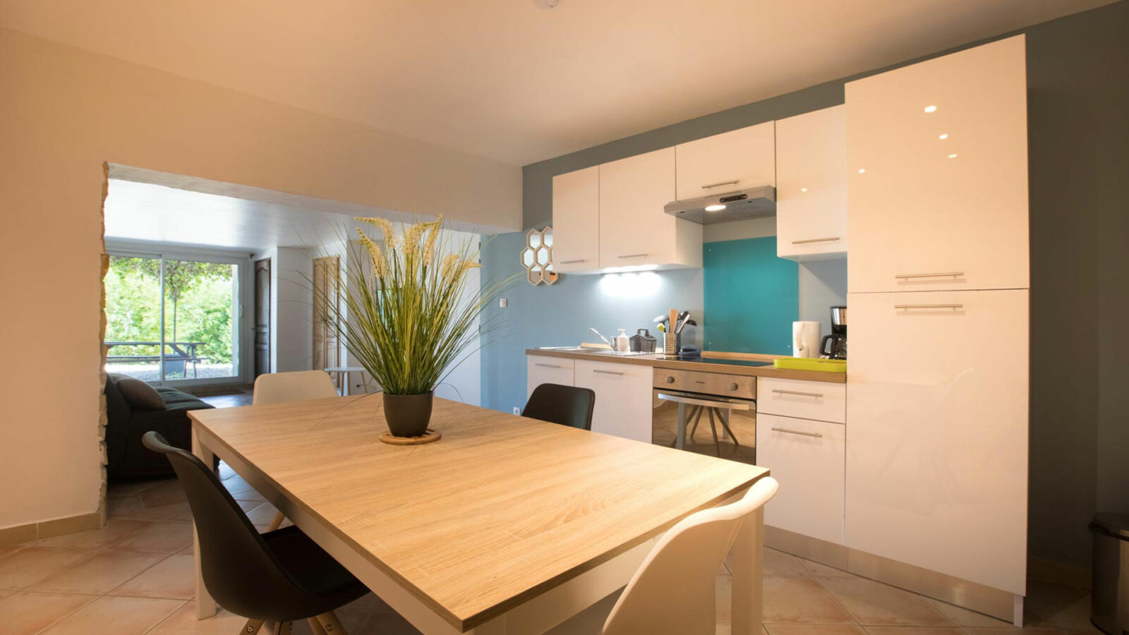 Modern kitchen with white cabinets, a wooden dining table, and four chairs—perfect for a groupe stay at a gîte Ardèche. A plant centerpiece brightens the table, while large windows and lighting fill the open space with natural light. Modern kitchen with white cabinets, a wooden dining table, and four chairs—perfect for a groupe stay at a gîte Ardèche. A plant centerpiece brightens the table, while large windows and lighting fill the open space with natural light.