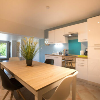 Modern kitchen with white cabinets, a wooden dining table, and four chairs—perfect for a groupe stay at a gîte Ardèche. A plant centerpiece brightens the table, while large windows and lighting fill the open space with natural light.