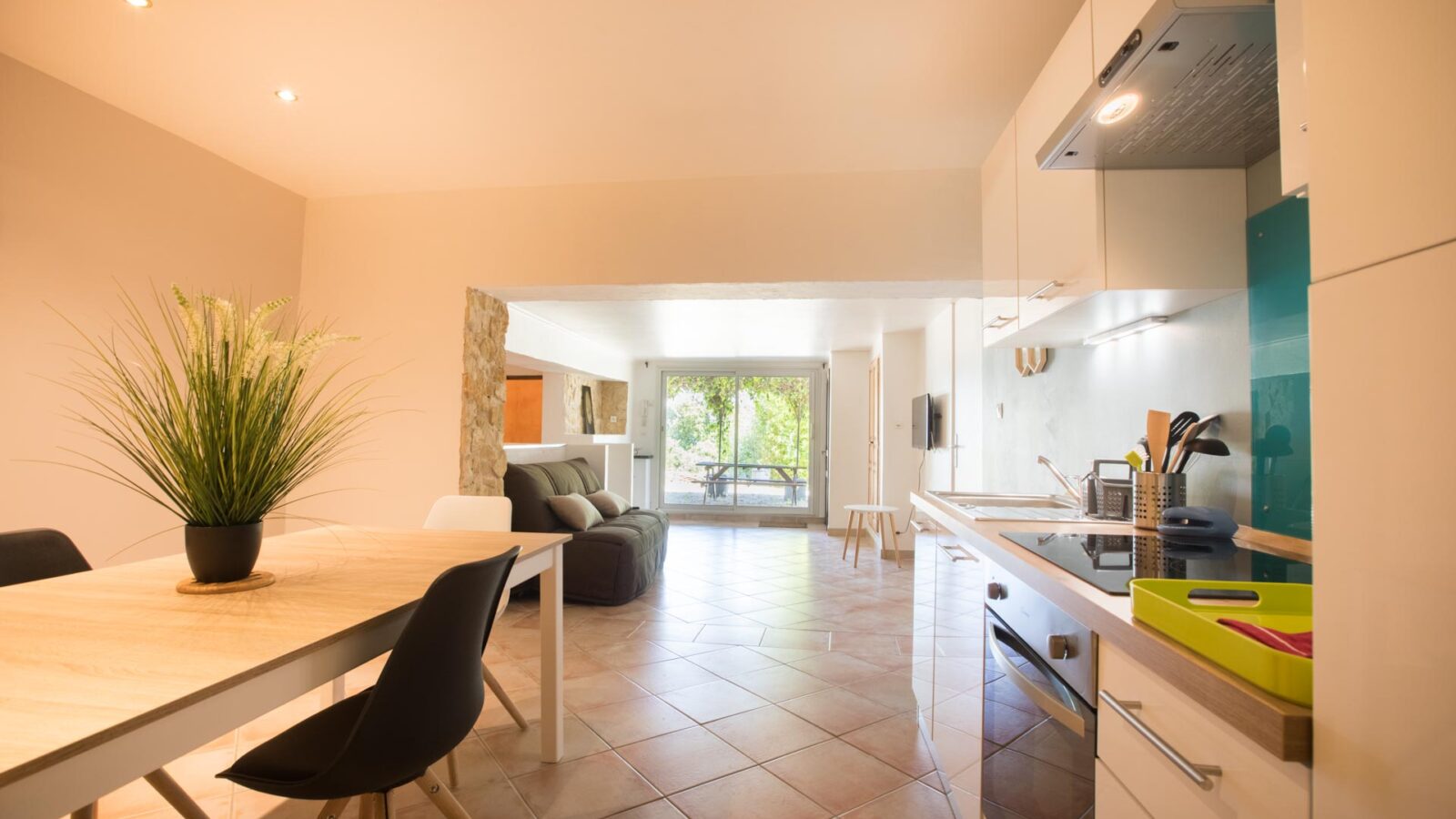 Modern kitchen and dining area with a wooden table, black and white chairs, potted plant, and appliances. This gîte Ardèche features an open layout leading to a bright living room with large windows and views of greenery outside. Modern kitchen and dining area with a wooden table, black and white chairs, potted plant, and appliances. This gîte Ardèche features an open layout leading to a bright living room with large windows and views of greenery outside.
