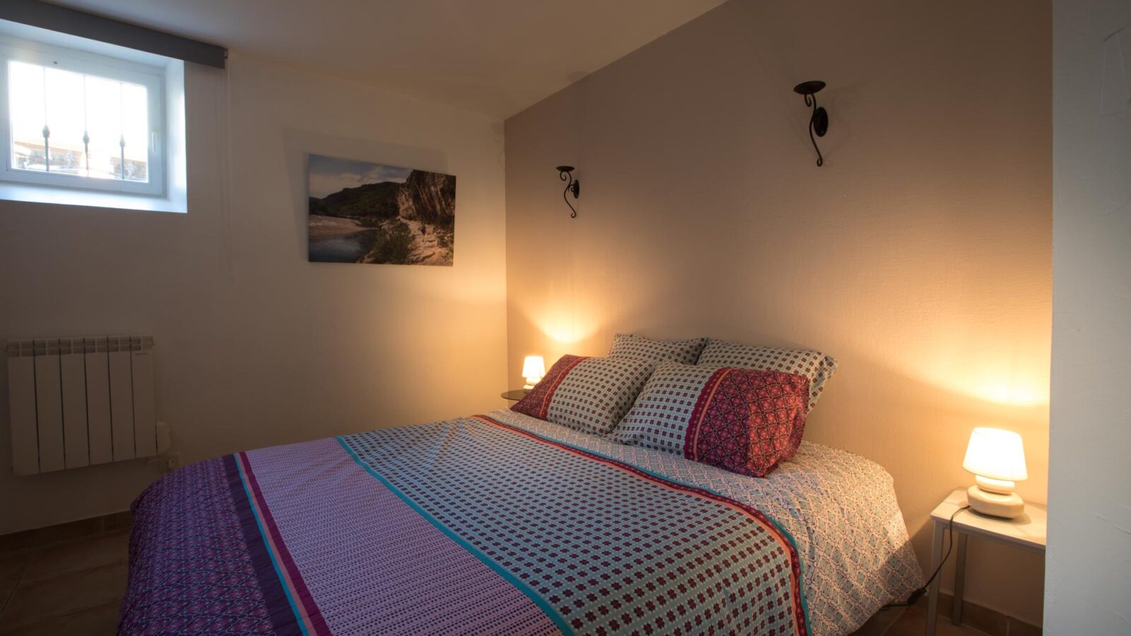 Cozy bedroom with a double bed covered in patterned bedding, two bedside tables with small lamps, wall-mounted lights, artwork on the wall, a small window letting in natural light—ideal for groupe stays in un hébergement Ardèche méridionale avec piscine. Cozy bedroom with a double bed covered in patterned bedding, two bedside tables with small lamps, wall-mounted lights, artwork on the wall, a small window letting in natural light—ideal for groupe stays in un hébergement Ardèche méridionale avec piscine.