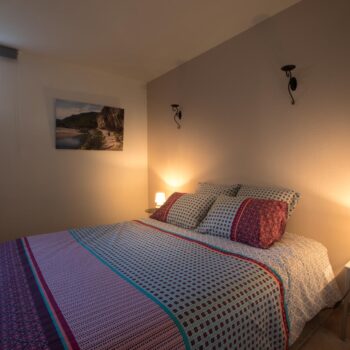 Cozy bedroom with a double bed covered in patterned bedding, two bedside tables with small lamps, wall-mounted lights, artwork on the wall, a small window letting in natural light—ideal for groupe stays in un hébergement Ardèche méridionale avec piscine.