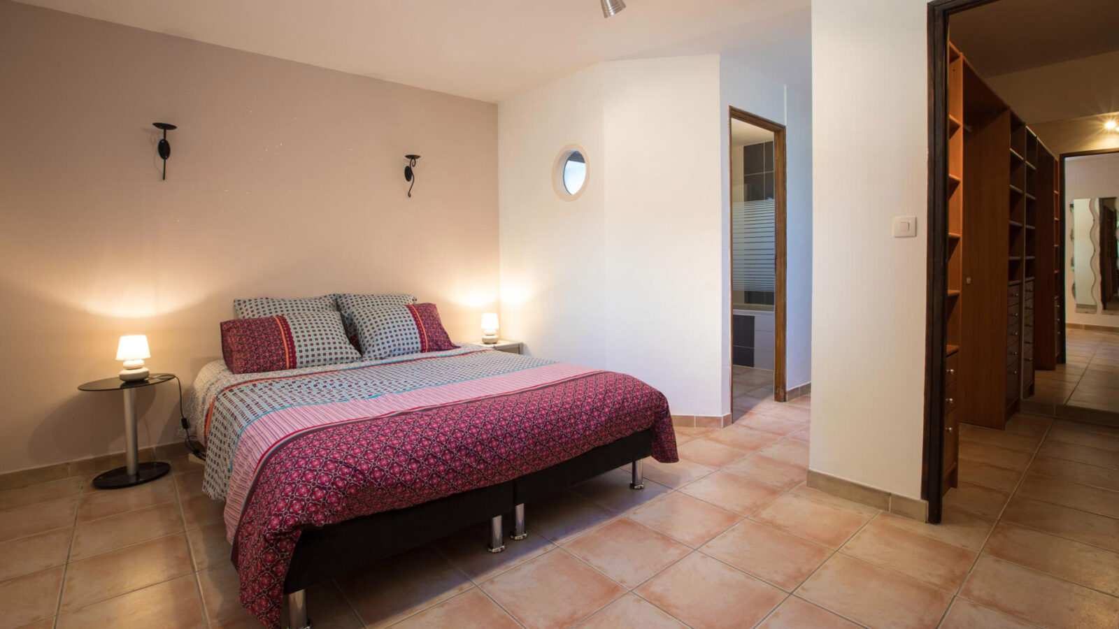 A tidy bedroom at Mas Saint Antoine gîte Ardèche features a double bed with patterned bedding, two side tables with lamps, tiled floor, neutral walls, and a hallway to a bathroom and walk-in closet—ideal for a groupe stay. A tidy bedroom at Mas Saint Antoine gîte Ardèche features a double bed with patterned bedding, two side tables with lamps, tiled floor, neutral walls, and a hallway to a bathroom and walk-in closet—ideal for a groupe stay.