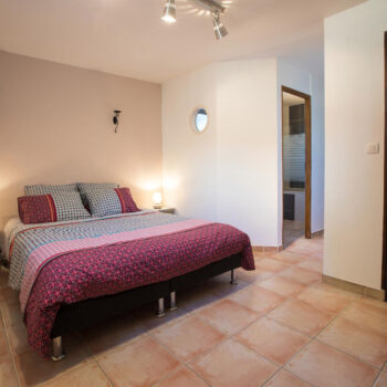 A tidy bedroom at Mas Saint Antoine gîte Ardèche features a double bed with patterned bedding, two side tables with lamps, tiled floor, neutral walls, and a hallway to a bathroom and walk-in closet—ideal for a groupe stay.