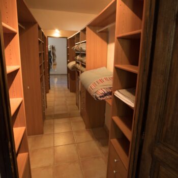 A narrow walk-in closet at Mas Saint Antoine, with wooden shelves on both sides holding folded blankets and linens, tiled floor, and a large mirror at the end reflecting the inviting space—perfect for groupe hébergement Ardèche méridionale.