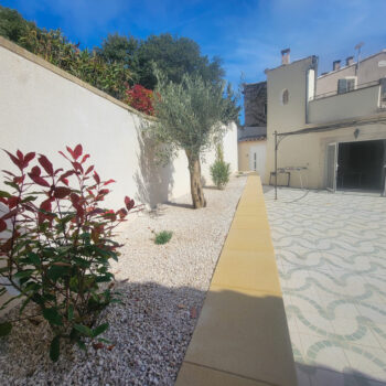 A sunny courtyard at a gîte Ardèche features patterned tile flooring, white gravel garden beds, small trees, and red-leaved shrubs—perfect for a groupe enjoying an authentic stay in the beautiful Ardèche méridionale.