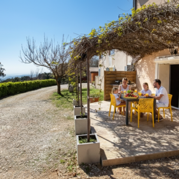 A family of four enjoys a meal together at a yellow table on a sunlit patio at Mas Saint Antoine, shaded by a vine-covered pergola, beside their gîte Ardèche with scenic countryside views and a dirt road nearby.
