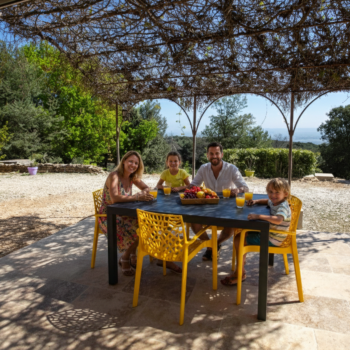 A family of four sits at a patio table under a vine-covered pergola at Mas Saint Antoine, enjoying drinks and fruit. They are outside in a sunny, rural setting with trees and distant hills, perfect for a relaxing gîte Ardèche escape.