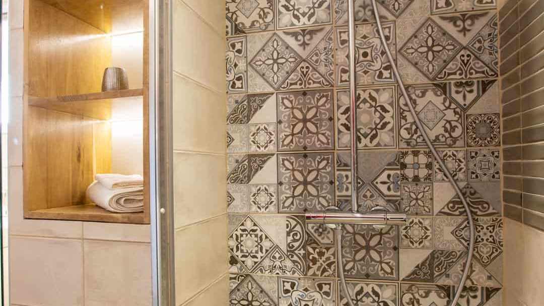 A modern shower at Mas Saint Antoine features patterned tiles, a rainfall showerhead, handheld shower, and a wooden shelf with rolled towels—perfect for your gîte Ardèche stay. The wood-style floor and ample lighting create a welcoming space for any groupe. A modern shower at Mas Saint Antoine features patterned tiles, a rainfall showerhead, handheld shower, and a wooden shelf with rolled towels—perfect for your gîte Ardèche stay. The wood-style floor and ample lighting create a welcoming space for any groupe.
