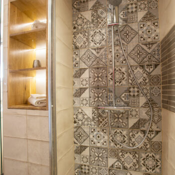 A modern shower at Mas Saint Antoine features patterned tiles, a rainfall showerhead, handheld shower, and a wooden shelf with rolled towels—perfect for your gîte Ardèche stay. The wood-style floor and ample lighting create a welcoming space for any groupe.