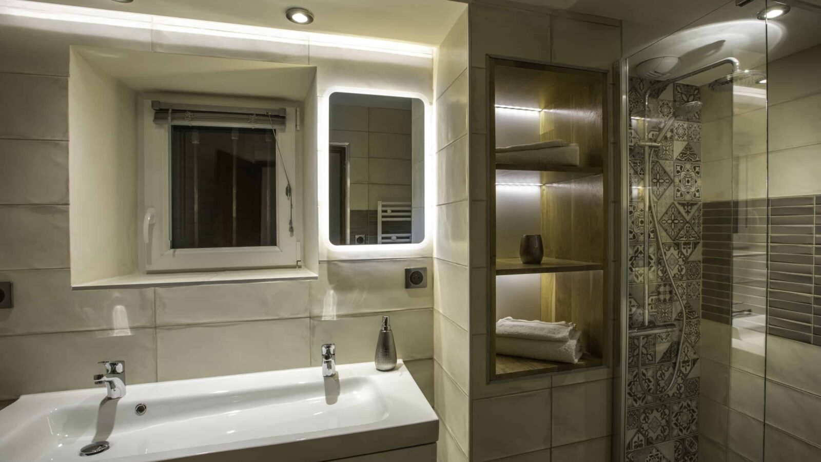 Modern bathroom in a gîte Ardèche with a white sink, illuminated mirror, built-in shelves for towels and decor, plus a glass shower with patterned tiles. Warm lighting creates a cozy retreat after enjoying the groupe’s day by the piscine. Modern bathroom in a gîte Ardèche with a white sink, illuminated mirror, built-in shelves for towels and decor, plus a glass shower with patterned tiles. Warm lighting creates a cozy retreat after enjoying the groupe’s day by the piscine.