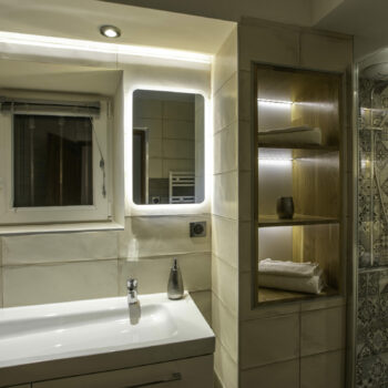 Modern bathroom in a gîte Ardèche with a white sink, illuminated mirror, built-in shelves for towels and decor, plus a glass shower with patterned tiles. Warm lighting creates a cozy retreat after enjoying the groupe’s day by the piscine.