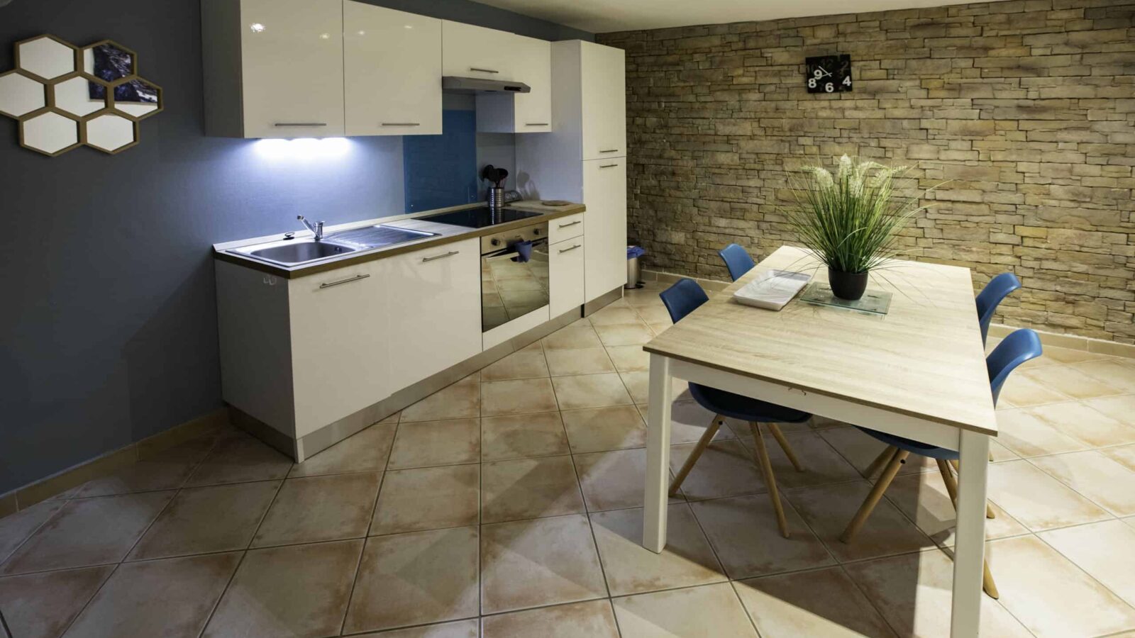 Modern kitchen with white cabinets, a sink, and stove along a blue accent wall. Perfect for a groupe getaway in this gîte Ardèche, it features a wooden dining table with blue chairs and a plant centerpiece near a stone accent wall. Modern kitchen with white cabinets, a sink, and stove along a blue accent wall. Perfect for a groupe getaway in this gîte Ardèche, it features a wooden dining table with blue chairs and a plant centerpiece near a stone accent wall.