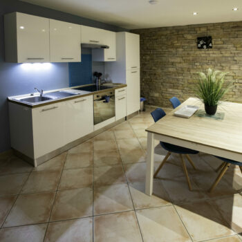 Modern kitchen with white cabinets, a sink, and stove along a blue accent wall. Perfect for a groupe getaway in this gîte Ardèche, it features a wooden dining table with blue chairs and a plant centerpiece near a stone accent wall.