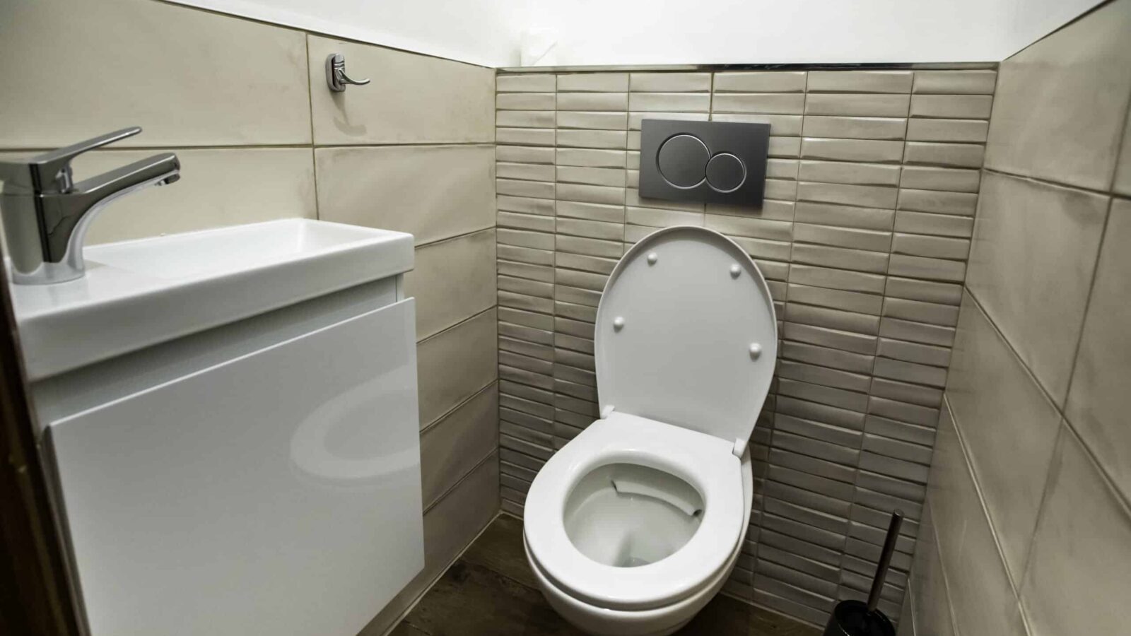 A small modern bathroom at Mas Saint Antoine with a white toilet, an open lid, a wall-mounted flush button, compact sink, tiled walls, and a toilet brush on a wooden floor—perfect for your groupe’s stay at the gîte Ardèche. A small modern bathroom at Mas Saint Antoine with a white toilet, an open lid, a wall-mounted flush button, compact sink, tiled walls, and a toilet brush on a wooden floor—perfect for your groupe’s stay at the gîte Ardèche.