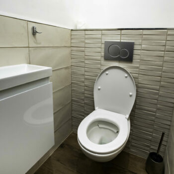 A small modern bathroom at Mas Saint Antoine with a white toilet, an open lid, a wall-mounted flush button, compact sink, tiled walls, and a toilet brush on a wooden floor—perfect for your groupe’s stay at the gîte Ardèche.