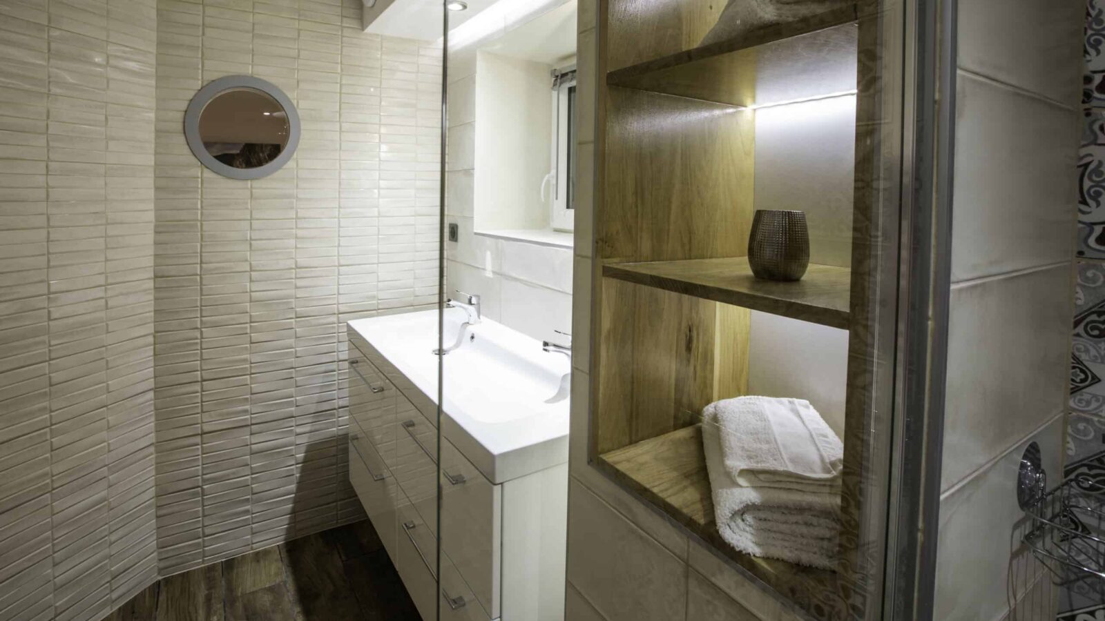 Modern bathroom at Mas Saint Antoine with beige textured tiles, a white sink vanity, round mirror, wooden shelves with towels and a vase, plus a glass shower door. Warm lighting adds comfort to your hébergement Ardèche méridionale experience. Modern bathroom at Mas Saint Antoine with beige textured tiles, a white sink vanity, round mirror, wooden shelves with towels and a vase, plus a glass shower door. Warm lighting adds comfort to your hébergement Ardèche méridionale experience.