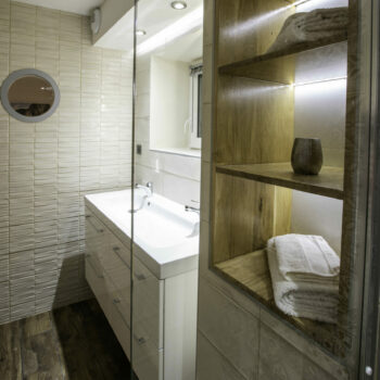 Modern bathroom at Mas Saint Antoine with beige textured tiles, a white sink vanity, round mirror, wooden shelves with towels and a vase, plus a glass shower door. Warm lighting adds comfort to your hébergement Ardèche méridionale experience.