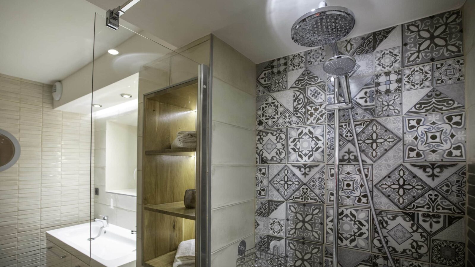 Modern bathroom at Mas Saint Antoine with a glass shower, patterned tile wall, large round showerhead, built-in wooden shelves with towels—perfect for groupe hébergement Ardèche méridionale—plus a white sink with mirror under soft lighting. Modern bathroom at Mas Saint Antoine with a glass shower, patterned tile wall, large round showerhead, built-in wooden shelves with towels—perfect for groupe hébergement Ardèche méridionale—plus a white sink with mirror under soft lighting.