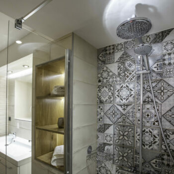 Modern bathroom at Mas Saint Antoine with a glass shower, patterned tile wall, large round showerhead, built-in wooden shelves with towels—perfect for groupe hébergement Ardèche méridionale—plus a white sink with mirror under soft lighting.